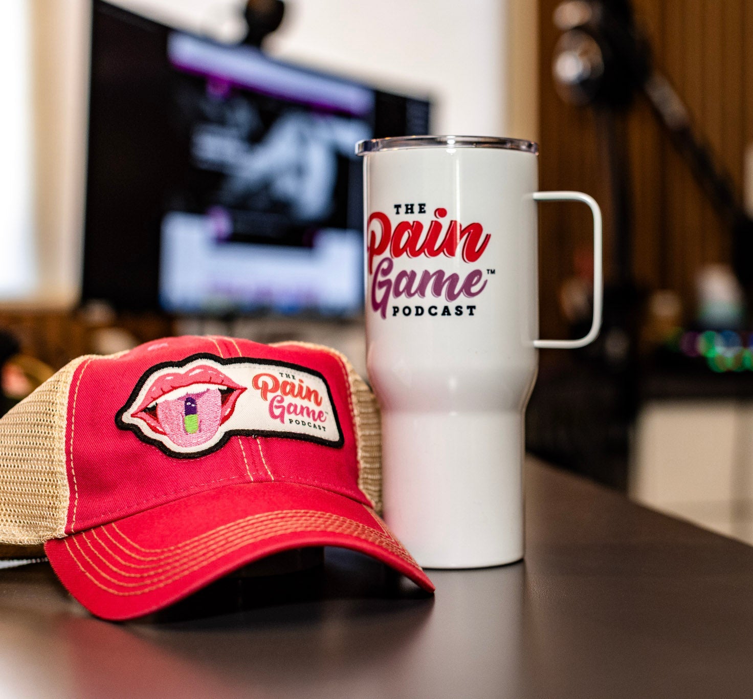 Red cap and white mug with 'The Pain Game Podcast' branding on a desk.